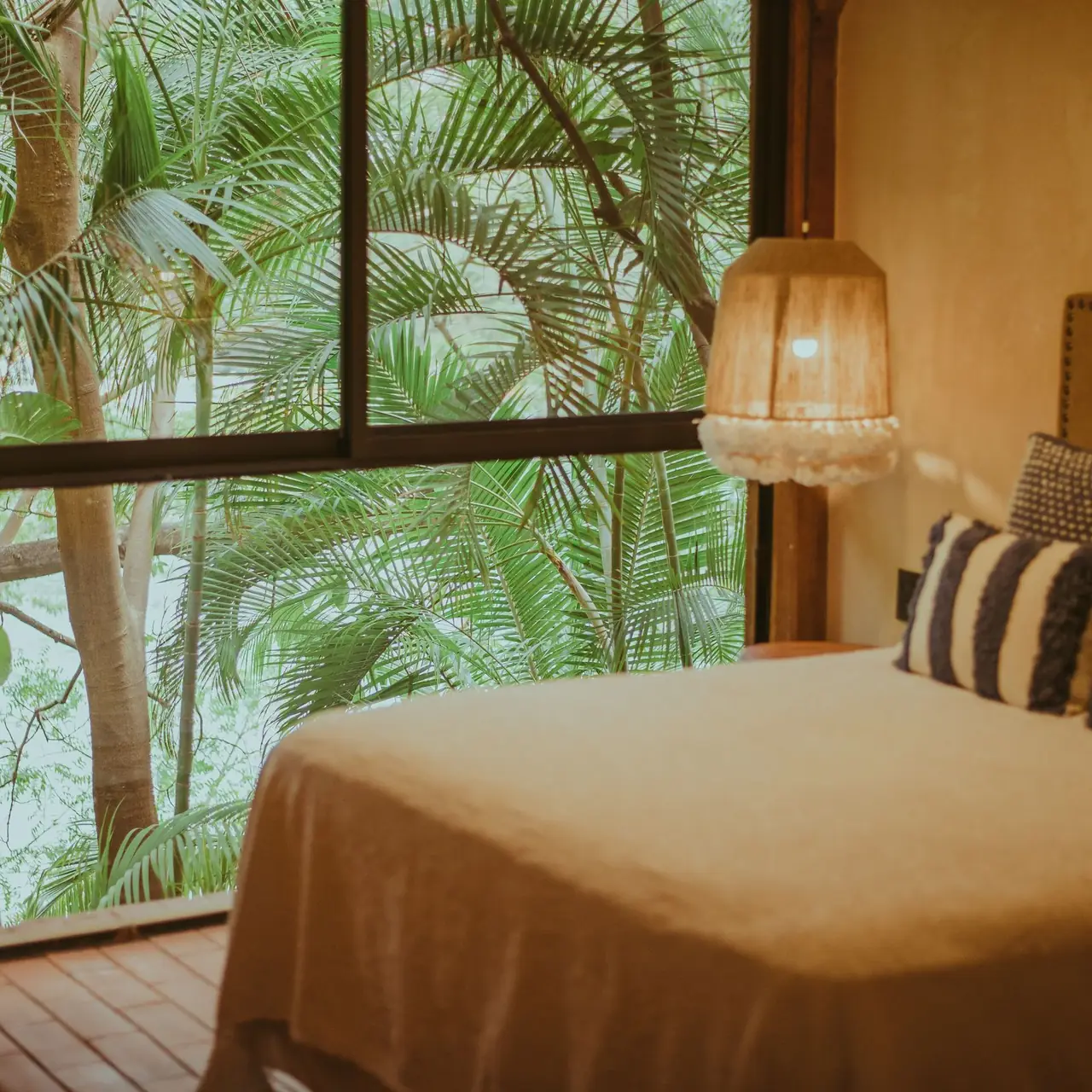A made bed beside a window looking out to palm leaves