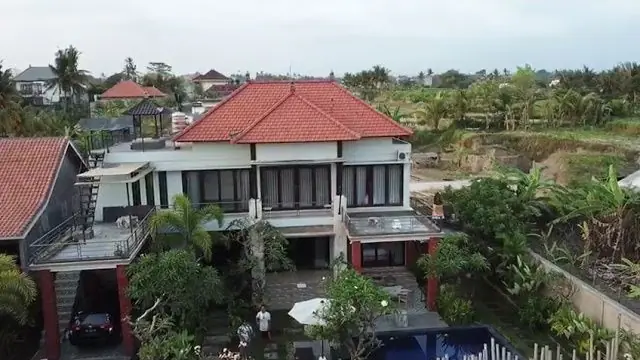 Drone view of the two-storey guest house with a red tiled roof, the pool and rice fields behind